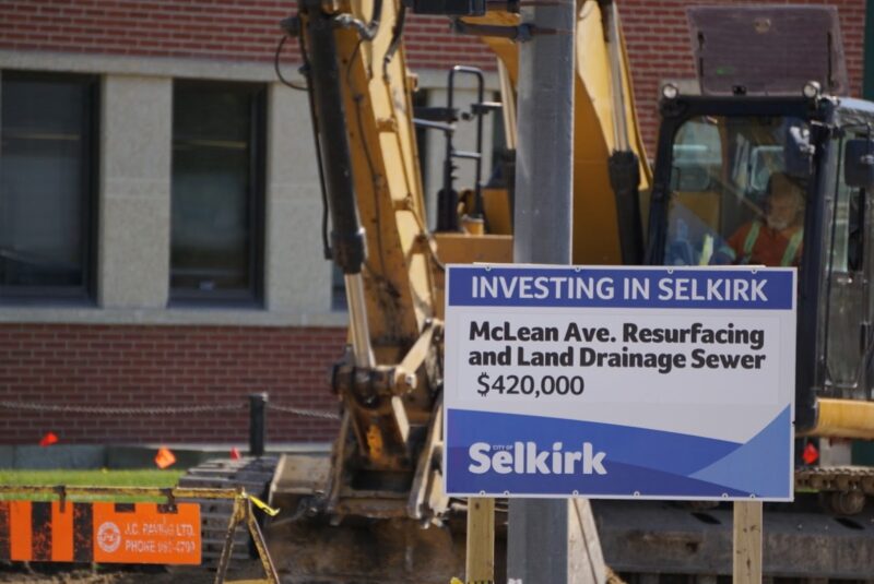 Sign saying "Investing in Selkirk McLean Ave. Resurfacing and Land Drainage Sewer $420,000" in front of an excavator