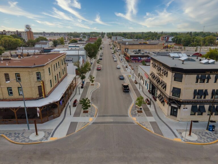 Aerial view of downtown Selkirk, Manitoba, looking down Manitoba Avenue East. Historic brick buildings with businesses line both sides of the street, including The Tractors Restaurant and a hotel with a patio. Cars are parked along the road, trees and planters line the sidewalks, and the sky is bright with scattered clouds.