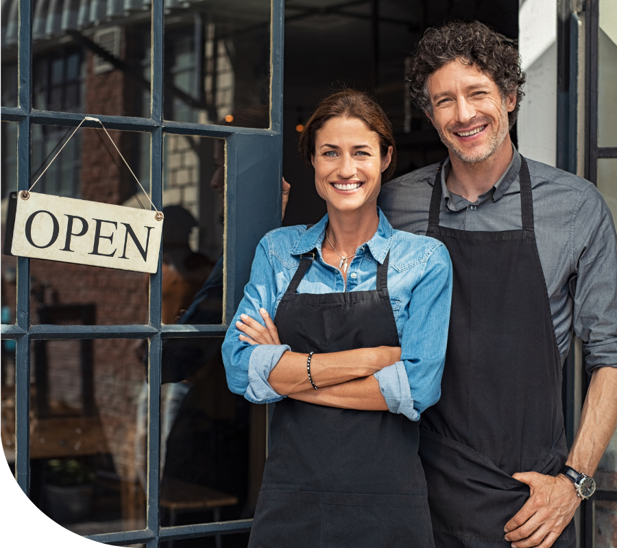 A female and male business owner standing outside their business displaying an "Open" sign.