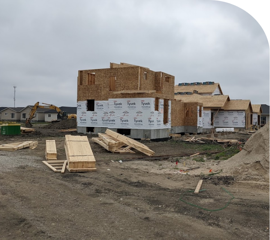 Residential development with a partially built house on muddy ground under a cloudy sky.